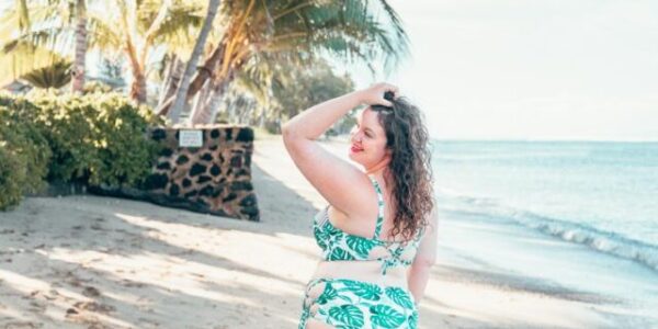 Curvy girl in a bikini printed with monstera leaves, turned away to face a beach with palm trees and water in the background.